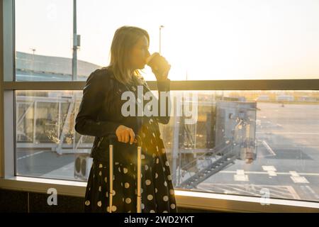 Pronta per salire a bordo con la sua valigia, una giovane donna tiene una tazza di caffè Foto Stock