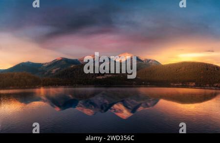 Splendido paesaggio con montagne vicino al lago con riflessi sull'acqua contro cielo nuvoloso blu e luce solare gialla al tramonto. Foto Stock