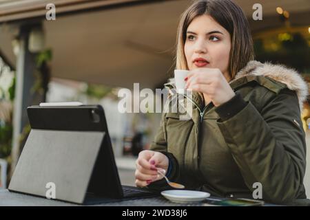 Una giovane donna curvida con una giacca verde si gode una tazza calda di caffè mentre lavora sul suo tablet, incarnando la flessibilità del lavoro remoto. Foto Stock