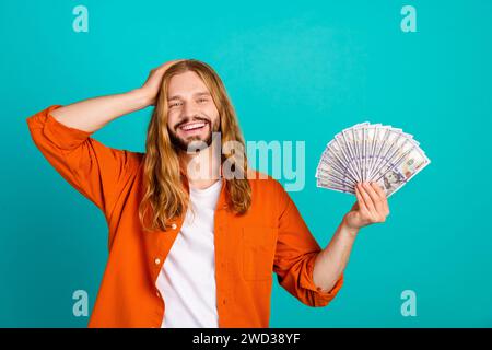 Foto di un allegro ragazzo con i capelli ondulati indossa un elegante outfit che tiene il braccio del denaro sulla testa isolato su sfondo turchese Foto Stock