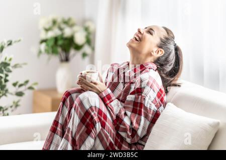 Giovane donna in pigiama rosso seduto sul divano e bevendo caffè al mattino in salotto, godendosi la giornata libera e ridendo. Foto Stock