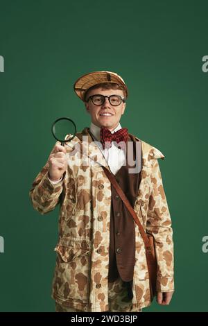 Giovane uomo che indossa abiti vintage, in piedi con una lente di ingrandimento sullo sfondo verde dello studio. La storia del detective Foto Stock