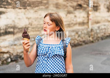 Simpatica bambina che mangia gelato al cioccolato all'aperto, indossa un abito e uno zaino blu Vichy, viaggia con i bambini Foto Stock