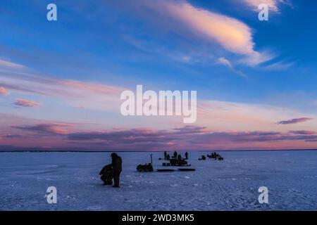 sagome di uomini pescatori e motoslitte durante la pesca invernale sul ghiaccio del fiume in serata contro il tramonto Foto Stock