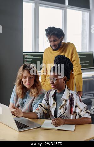 Colleghi birazziali seduti alla scrivania che guardano lo schermo del notebook, il loro collega indiano in piedi dietro Foto Stock