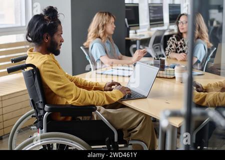 Giovane indiano in sedia a rotelle che lavora su un computer portatile seduto al tavolo dell'ufficio con i suoi colleghi Foto Stock