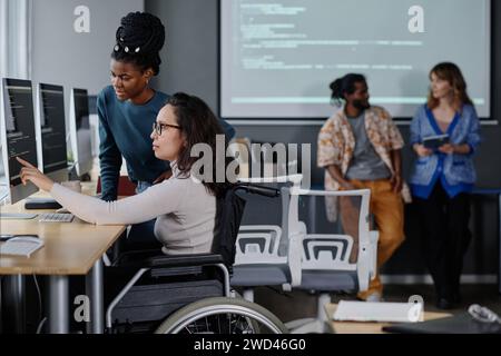 Donna nera che aiuta il suo collega in sedia a rotelle con la scrittura di codice, colleghi in piedi sullo sfondo Foto Stock