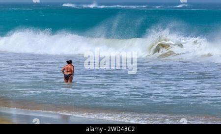Boa Vista, Capo Verde - 22 marzo 2018: Una grande onda con acqua turchese e schiuma bianca si avvicina ad una donna che rimane sulla spiaggia in una giornata ventosa . Foto Stock