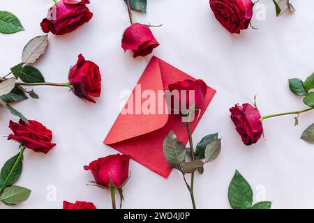 Due rose rosse e una lettera in una busta collocata su uno sfondo di marmo bianco immacolato, con una spruzzata di carta rossa Foto Stock