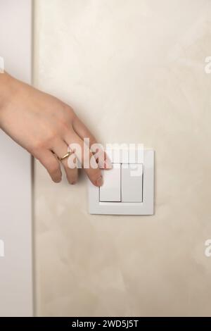 Il dito di una donna accende l'interruttore della luce a casa. Primo piano Una donna accende la luce con un interruttore a muro con la mano. Concentrati sull'interruttore Foto Stock