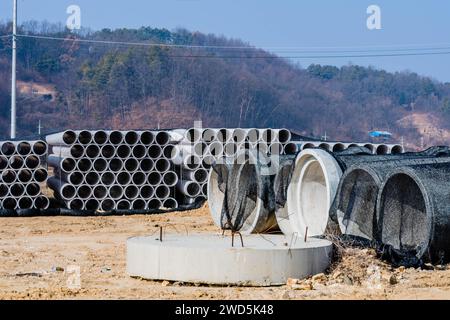 Grandi tombini scatolari rotondi di cemento davanti a pile di tubi ondulati rivestiti in rete nera in cantiere con alberi ricoperti di montagna Foto Stock