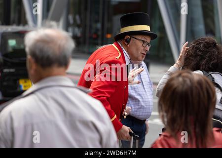 Un concierge vestito in elegante uniforme rossa tradizionale all'esterno del Rubens Hotel a Victoria, nel centro di Londra. Sta aiutando un passeggero a salire su un taxi nero Foto Stock