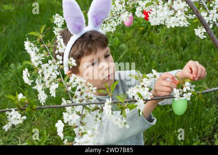 Caccia all'uovo di Pasqua. Bambino in età prescolare che indossa orecchie da coniglio per raccogliere uova colorate durante la caccia all'uovo di Pasqua in giardino. rami di decorazione per bambini dell'albero fiorito w Foto Stock