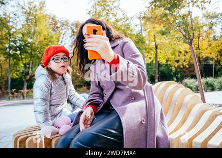 Bruna giovane madre che mostra cartoni animati a disabili bambina adorabile sullo smartphone. Donna che registra un video di famiglia con una figlia piccola in età prescolare accesa Foto Stock