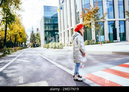 Vista laterale foto a tutta altezza di una graziosa bambina con un giocattolo che indossa un cappotto e un berretto rosso che si erge da solo vicino alla passeggiata sulla strada della città. Foto Stock