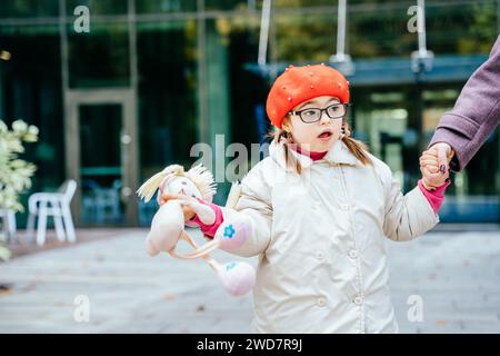 Simpatica ragazza con occhiali da vista che indossa berretto rosso cappotto con esigenze speciali che tiene la mano della madre durante una passeggiata all'aperto. Foto Stock