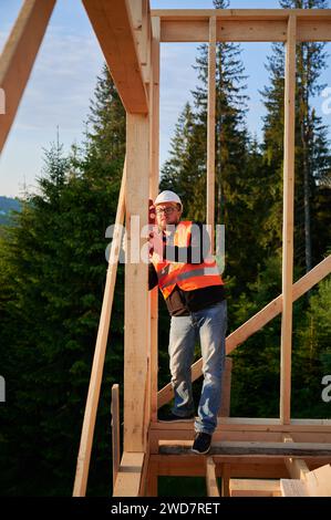 Falegname che costruisce case con telaio in legno. L'uomo barbuto, indossando occhiali, ispeziona le pareti per la livellatezza usando la livella a bolla d'aria. Concetto di costruzione contemporanea ecocompatibile. Foto Stock