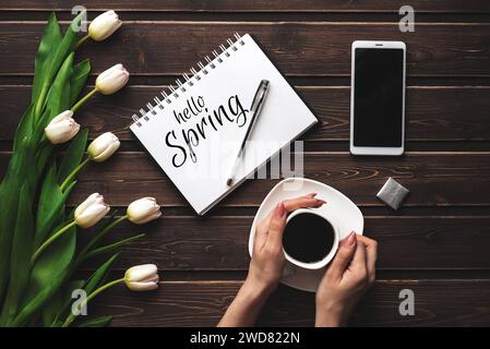 Biglietto d'auguri con la scritta Hello Spring. Bouquet di tulipani bianchi e una tazza di caffè con uno smartphone su uno sfondo di legno Foto Stock