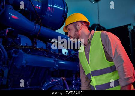 un tecnico dall'aspetto elegante e attento con giubbotto di sicurezza con casco e barba che lavora con attenzione, dati Foto Stock