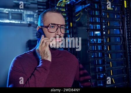 bellissimo specialista concentrato con occhiali e barba che parla al telefono al lavoro, nel data center Foto Stock
