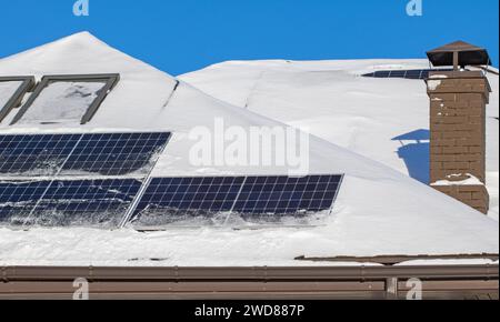 Un'immagine ad alto angolo di un tetto residenziale con pannelli solari puliti dalla neve, che fornisce energia ecologica sotto un cielo blu brillante durante il mare invernale Foto Stock