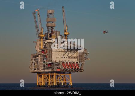 Elicottero che vola verso la piattaforma petrolifera nell'oceano per atterrare, in una giornata di sole limpido, con cielo colorato e mare blu calmo. Foto Stock