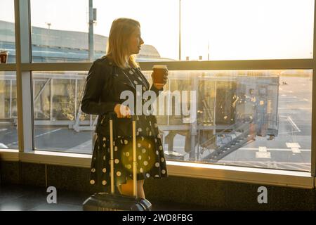 Una giovane donna che tiene una tazza di caffè è pronta per salire a bordo con la sua valigia. Foto Stock