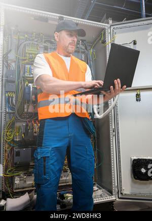 un elettricista in un cappuccio e un giubbotto arancione installa una grande sottostazione elettrica utilizzando un computer. Foto Stock