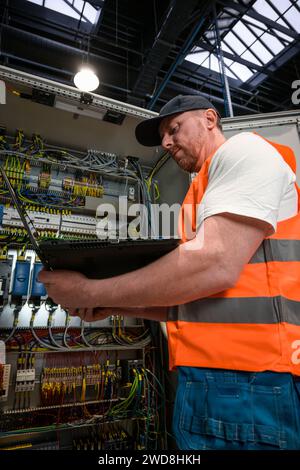 un elettricista in un cappuccio e un giubbotto arancione installa una grande sottostazione elettrica utilizzando un computer. Foto Stock