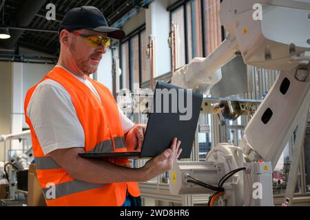 un ingegnere in un gilet sta programmando un robot su una linea di assemblaggio utilizzando un computer portatile. Foto Stock