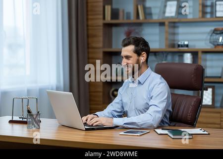 Un professionista maschio sorridente esperto che si occupa di affari tramite un computer portatile presso il suo ufficio domestico, mostrando il lavoro remoto. Foto Stock