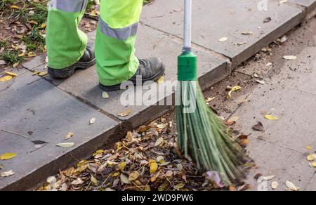 Un lavoratore municipale spazza la strada con il bastone di scopa e raccoglie i rifiuti in una paletta. operaio sanitario spazzatura strada Foto Stock