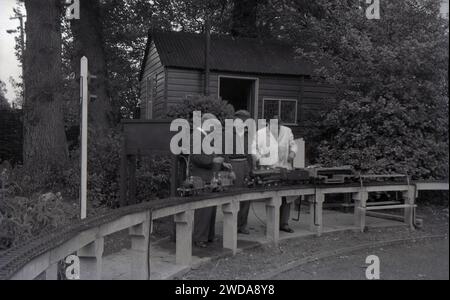 Anni '1950, storico, all'aperto in un giardino, appassionati di modellini di ferrovie , uno con un mantello bianco, accanto a una ferrovia a vapore in giardino in miniatura, presso la Field End Railway Co, Inghilterra, Regno Unito. Su un binario sopraelevato seduto su pali di cemento, il modello di treni a vapore, uno dei quali è un modello in scala della locomotiva a vapore GWR 'County of Oxford' (1023), il cui originale iniziò il servizio nel 1947. Foto Stock