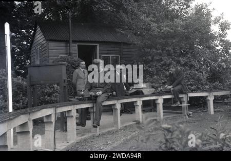 Anni '1950, storico, all'aperto in un giardino, appassionati di modellismo ferroviario a vapore in un giardino in miniatura, presso la Field End Railway Co, Inghilterra, Regno Unito. Su un binario sopraelevato seduto su pali di cemento, un uomo in giacca e berretto che cavalca un modellino di treno a vapore. Foto Stock