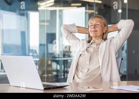 Una donna d'affari dai capelli grigi è seduta in ufficio alla scrivania con gli occhi chiusi, le mani dietro la testa e riposa. Foto Stock