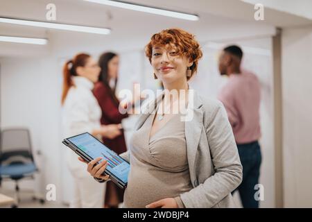 Donna d'affari creativa e incinta in posa con il laptop mentre i suoi compagni di squadra lavorano dietro per aumentare i profitti e la produttività in un piccolo ufficio. Foto Stock
