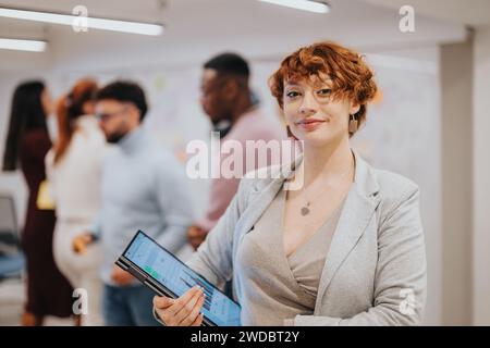 Splendida donna d'affari incinta che tiene un computer portatile in ufficio mentre il suo team sta facendo brainstorming e scrivendo su una parete di vetro sul retro. Copia spazio Foto Stock