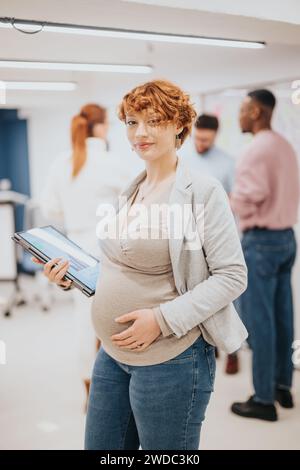 Splendida donna d'affari incinta rossa che si posa in ufficio mentre tiene il laptop. La sua squadra sta facendo il brainstorming alle spalle. Copia spazio. Foto Stock