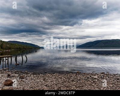 Un lago tranquillo con una spiaggia di ciottoli e un molo sotto un cielo nuvoloso, ai margini di Loch Ness. Scozia, Gran Bretagna Foto Stock