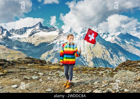 Graziosa bambina che indossa un cappotto colorato con arcobaleno brillante, che regge la bandiera svizzera, in piedi di fronte al ghiacciaio del Gornergrat, Svizzera Foto Stock