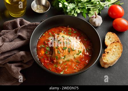 Pollo al forno con sugo di pomodoro ed erbe aromatiche in un recipiente su fondo di pietra scura. Gustoso stufato di pollo fatto in casa per cena. Vista ravvicinata Foto Stock