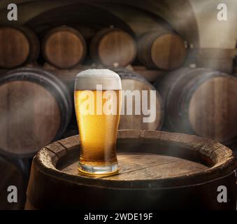 Bicchiere di birra in cima a botte di legno e botti di legno in cantina sullo sfondo. Raggi solari che cadono sulla superficie. Posizionamento del prodotto. Foto Stock