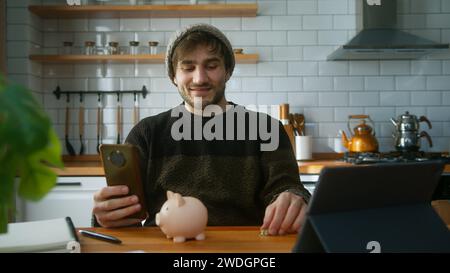 Giovane con berretto seduto in una cucina moderna a casa mentre tiene lo smartphone in mano guardando la fotocamera e mette un paio di monete nella p Foto Stock