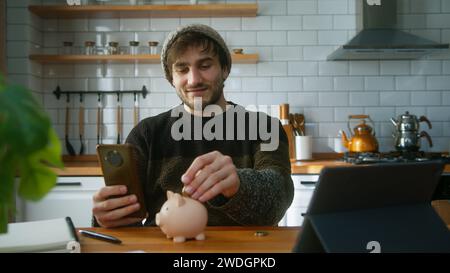 Uomo sorridente con berretto seduto in una cucina moderna a casa mentre tiene lo smartphone in mano guardando la fotocamera e mette un paio di monete nel Foto Stock