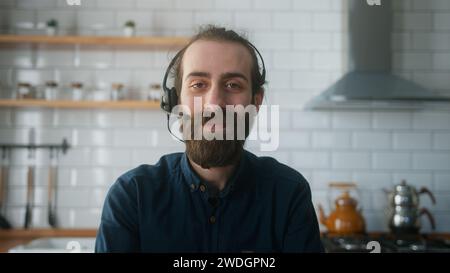 Un giovane uomo barbuto con un panino per capelli seduto in cucina a casa indossa cuffie wireless con microfono che parla con la macchina fotografica e conduce conferenze in bus Foto Stock