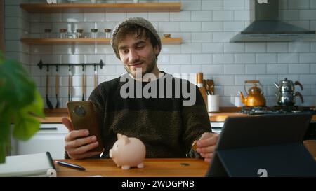 Uomo felice con berretto seduto in una cucina moderna a casa mentre tiene lo smartphone in mano guardando la fotocamera e mette un paio di monete nel pi Foto Stock