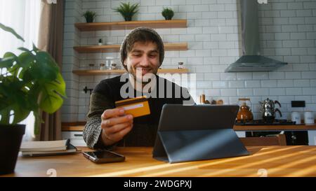 Uomo sorridente con berretto seduto in cucina a casa inserendo il numero di carta di credito sul tablet per rendere sicuro e facile il pagamento elettronico a distanza Foto Stock