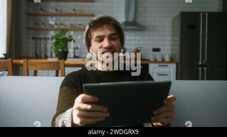 Un giovane con un berretto seduto sul divano a casa con lo sfondo della cucina, giocando a un eccitante concentrato impegnato in un interessante gioco mobil con ta Foto Stock