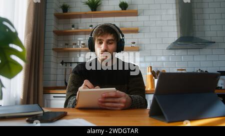 Bell'uomo con berretto seduto in una cucina moderna a casa, che indossa cuffie per ascoltare musica mentre lavora con il suo computer tablet e prende appunti Foto Stock