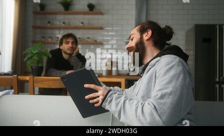 Un uomo barbuto seduto sul divano che parla con il suo amico seduto in cucina a casa, mostrando qualcosa sul suo tablet Foto Stock